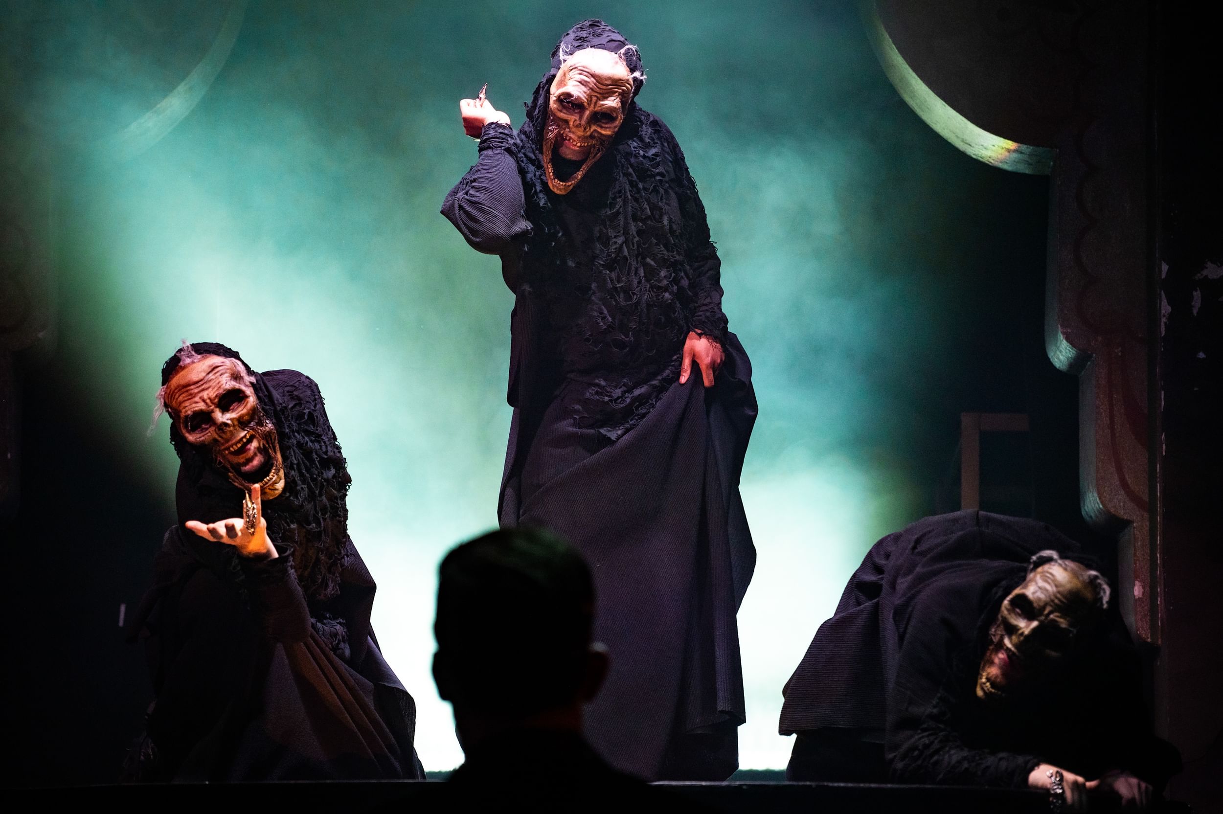 Against a dramatic green mist, three black-draped witches in creepy skull-like masks strike contorted positions.