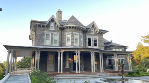 A Victorian-era stone mansion with peaked roof features and a large porch extending on its front and left side.