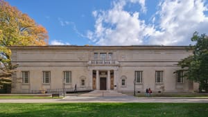 Exterior front view of the building’s pale, rectangular face, w/ tall symmetrical windows, pillared entrance & elegant stairs