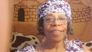 Portrait snapshot of Ongoza, a Black woman, wears matching white floral top and head wrap against a cloth backdrop