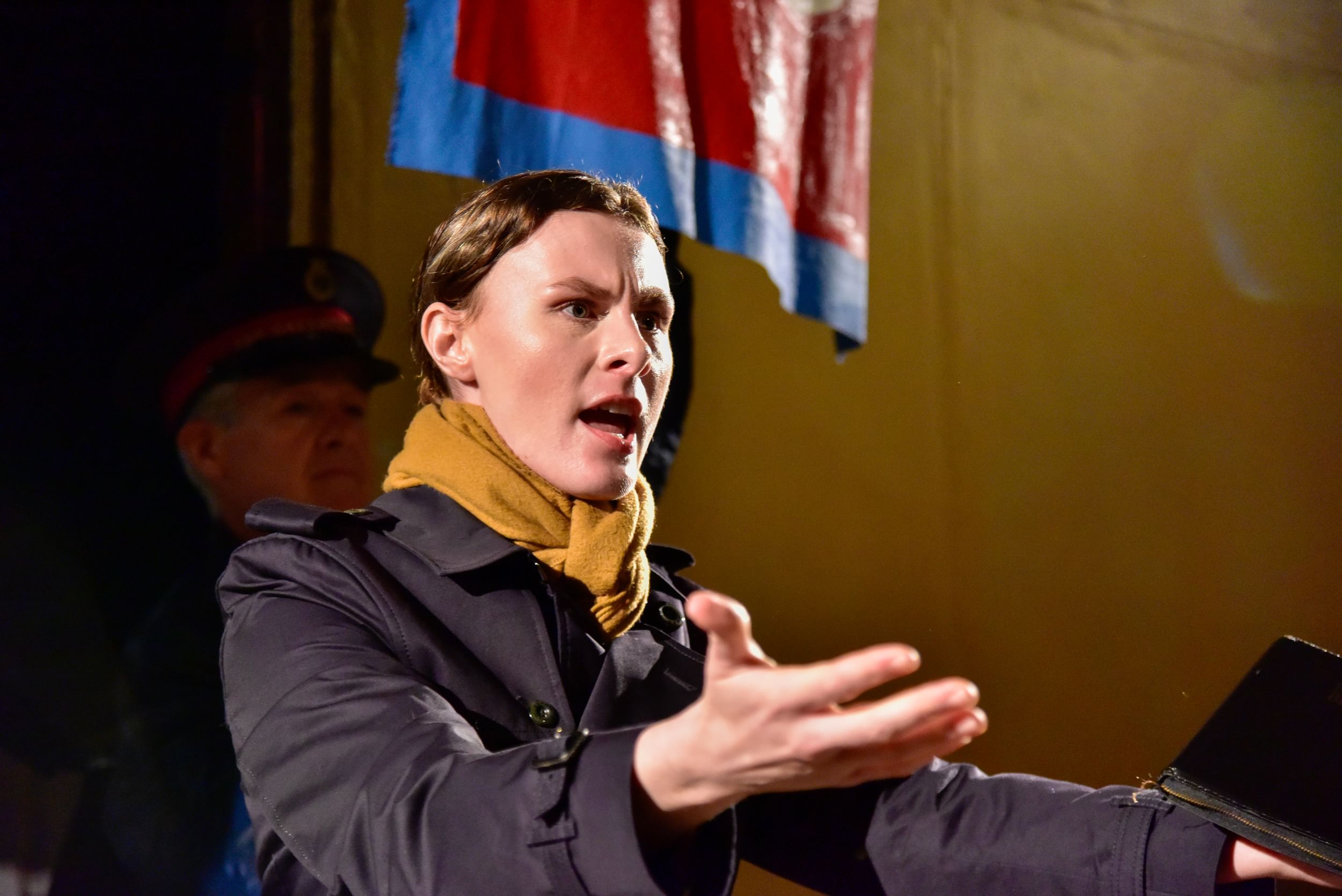 With a severe, animated expression, Ladd speaks and stretches out her arms, wearing a navy jacket and yellow scarf.