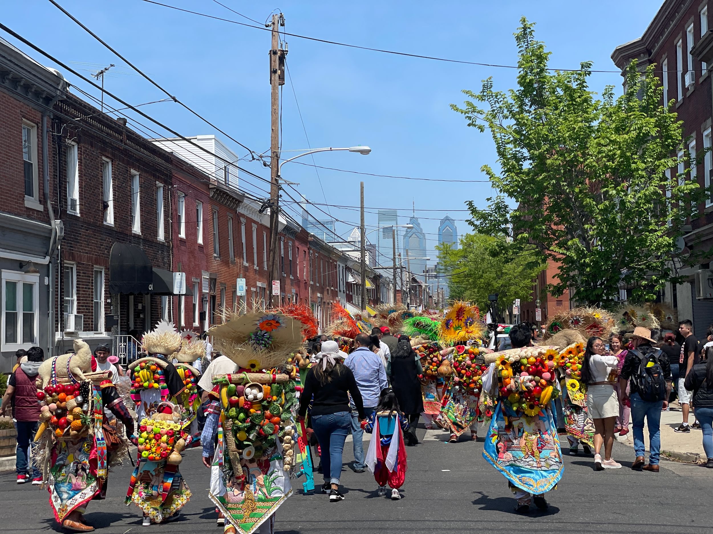 Seen from behind, a festival parade of marchers under the Philly skyline, in elaborate applique costumes with fruit & veggies