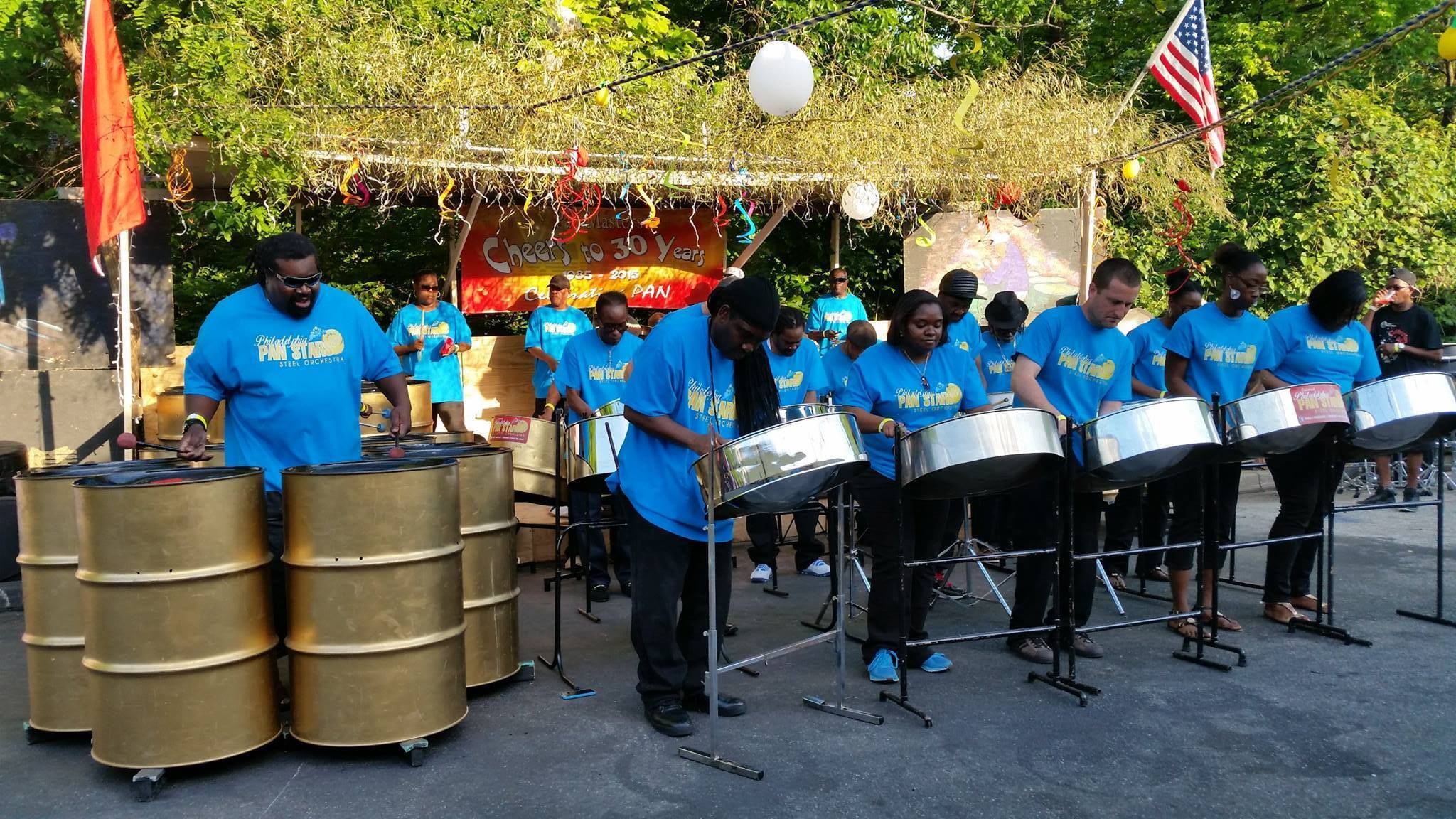 About 15 percussionists in blue shirts playing different gold and silver drums outside on a sunny day.