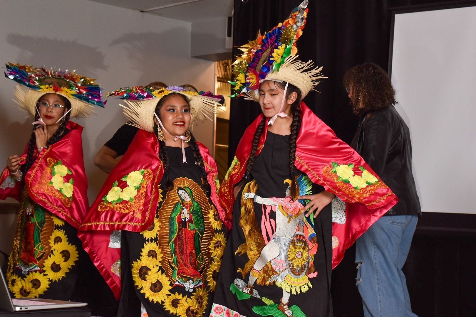 Three Latina girls wear elaborate costumes with bright religious & Indigenous applique designs & lavish decorated straw hats
