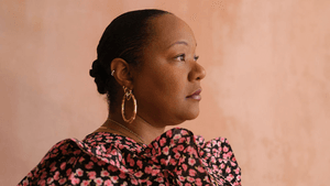 Holmes, a Black woman, in a layered floral black top & hoop earrings. She looks to the right against a melon-colored backdrop