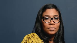 Rodgers, a Black woman. Portrait style photo with her to the right, glasses, and a yellow sweater over a black top
