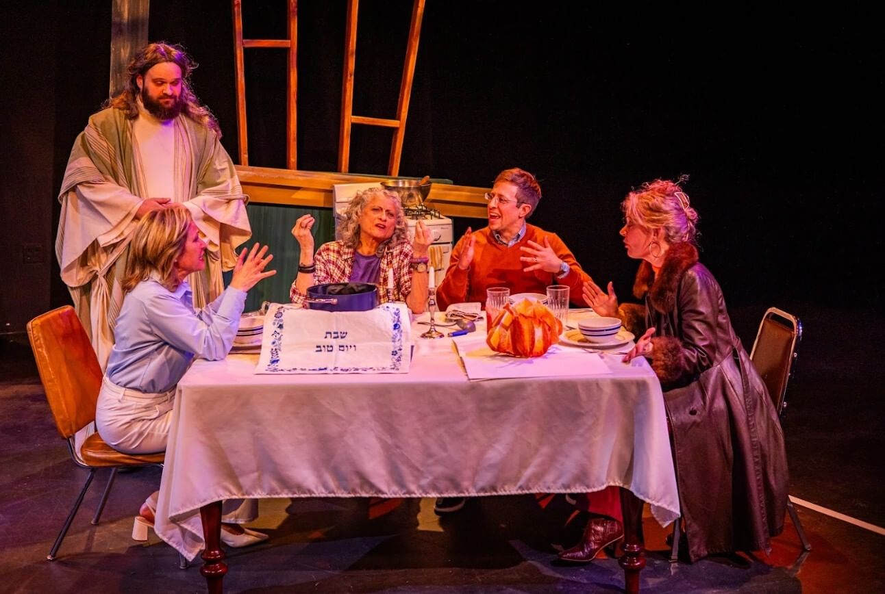 Four actors appear to argue animatedly while sitting at festive Jewish board. Another, evoking Jesus, stands calmly at left