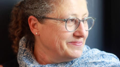 Portrait photo of Toll. She has gray hair pulled back in a pony tail, glasses, and a blue and white floral scarf.