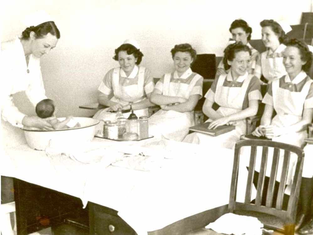 Vintage black & white photo of a woman bathing an infant in a basin while six smiling nurses in uniform look on smiling