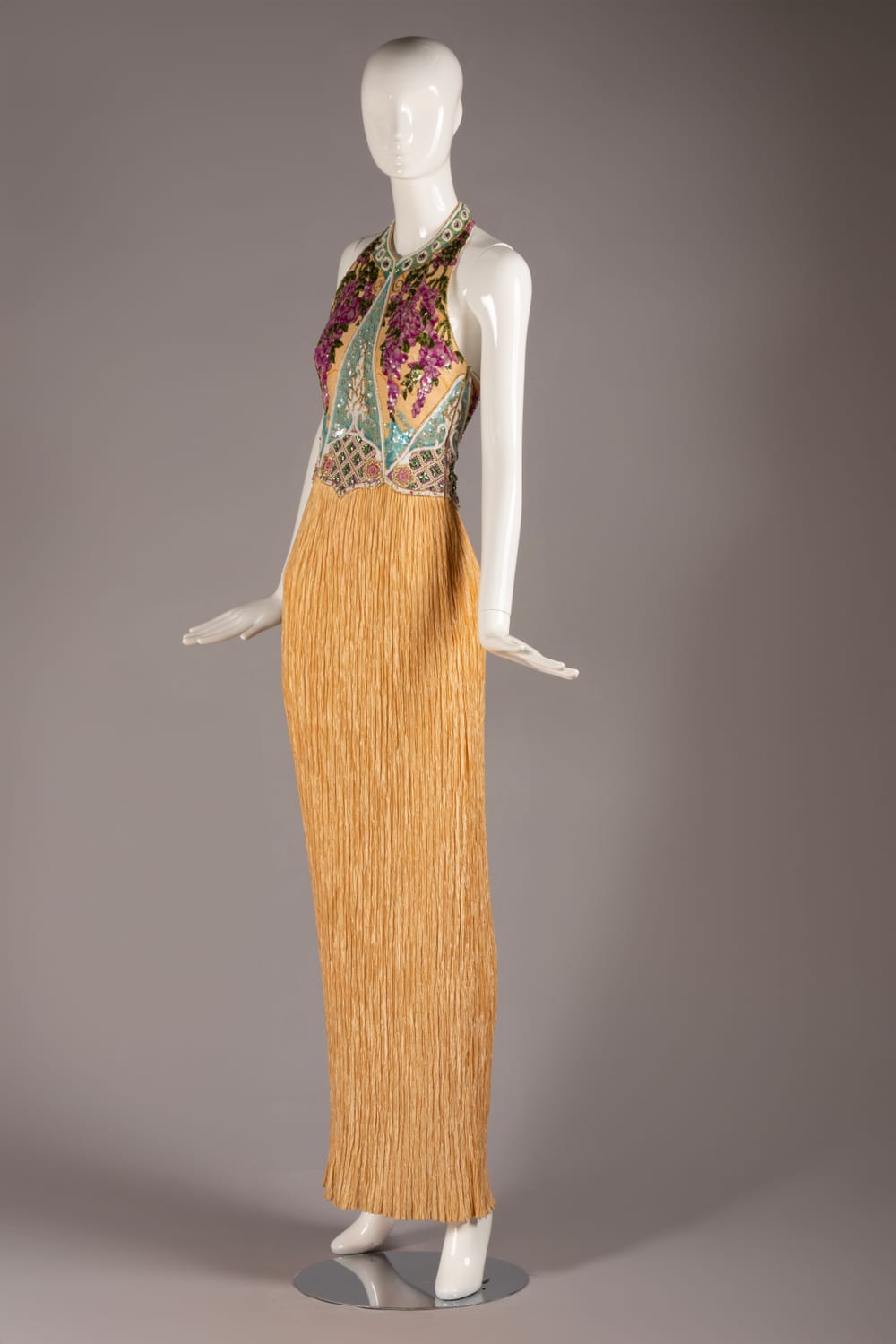 A gold, slender halter-style dress with a long, delicate pleated skirt and opulent beaded bodice in muted purple and blue.