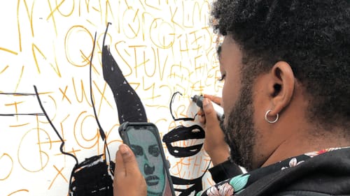 Close-up over Mcilwaine’s shoulder as he draws Lady’s Liberty’s features on the wall with a fat black marker.