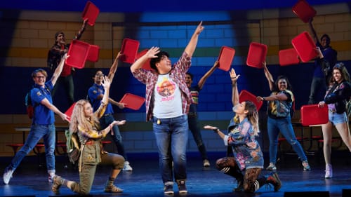 In the middle of the cast holding red lunch trays like fans, Jih-Cook poses joyfully in jeans & plaid, pointing one hand up