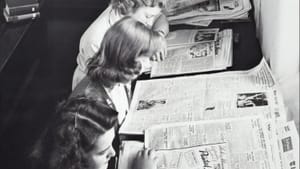 Black & white vintage photo of five coiffed young ladies sitting in a row in the library reading newspapers.