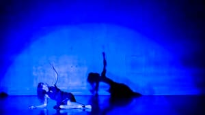 A dancer on stage, tinted by the deep blue light cast over her on stage, her shadow cast large on the wall behind her