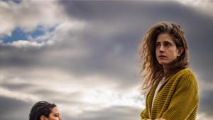 Two women stand on large rocks near a river or shore, gray skies. They look on in opposite directions, ominous, concerned