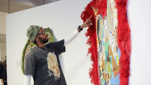 Melancon, in black tee, glasses, and a camo hat, gestures to his large glass bead piece on the wall, rimmed with red feathers