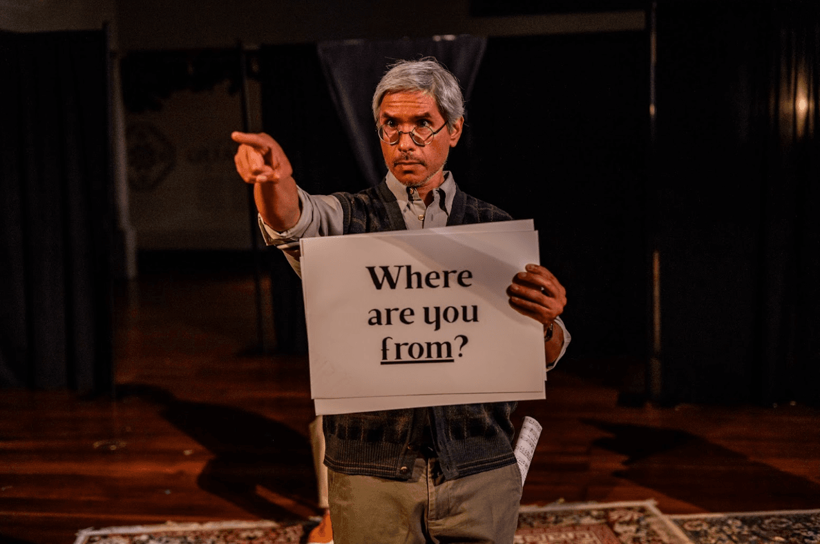 Hernandez, with silver hair & round glasses, holds a white sign reading “Where are you from?” and points seriously outward.