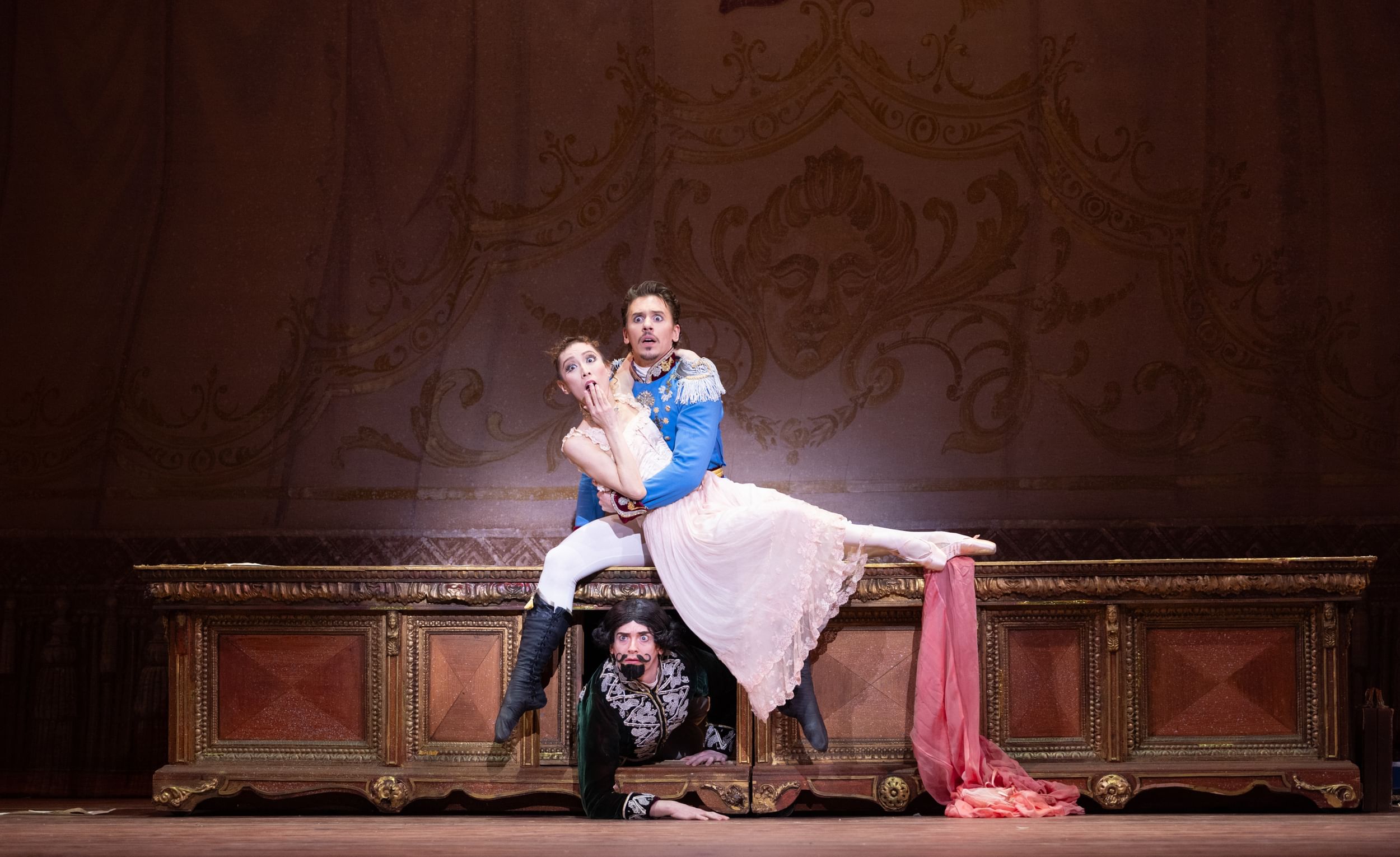 Iseda & Roxander, horrified as their tryst is interrupted, sit on an ornate desk while the mustached Drucker crawls out