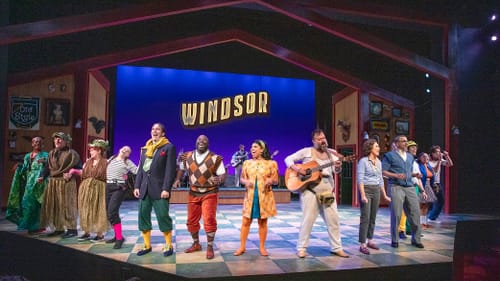 14 cast members, in colorful eclectic costumes, line up joyously for a curtain call at the curving edge of the stage.