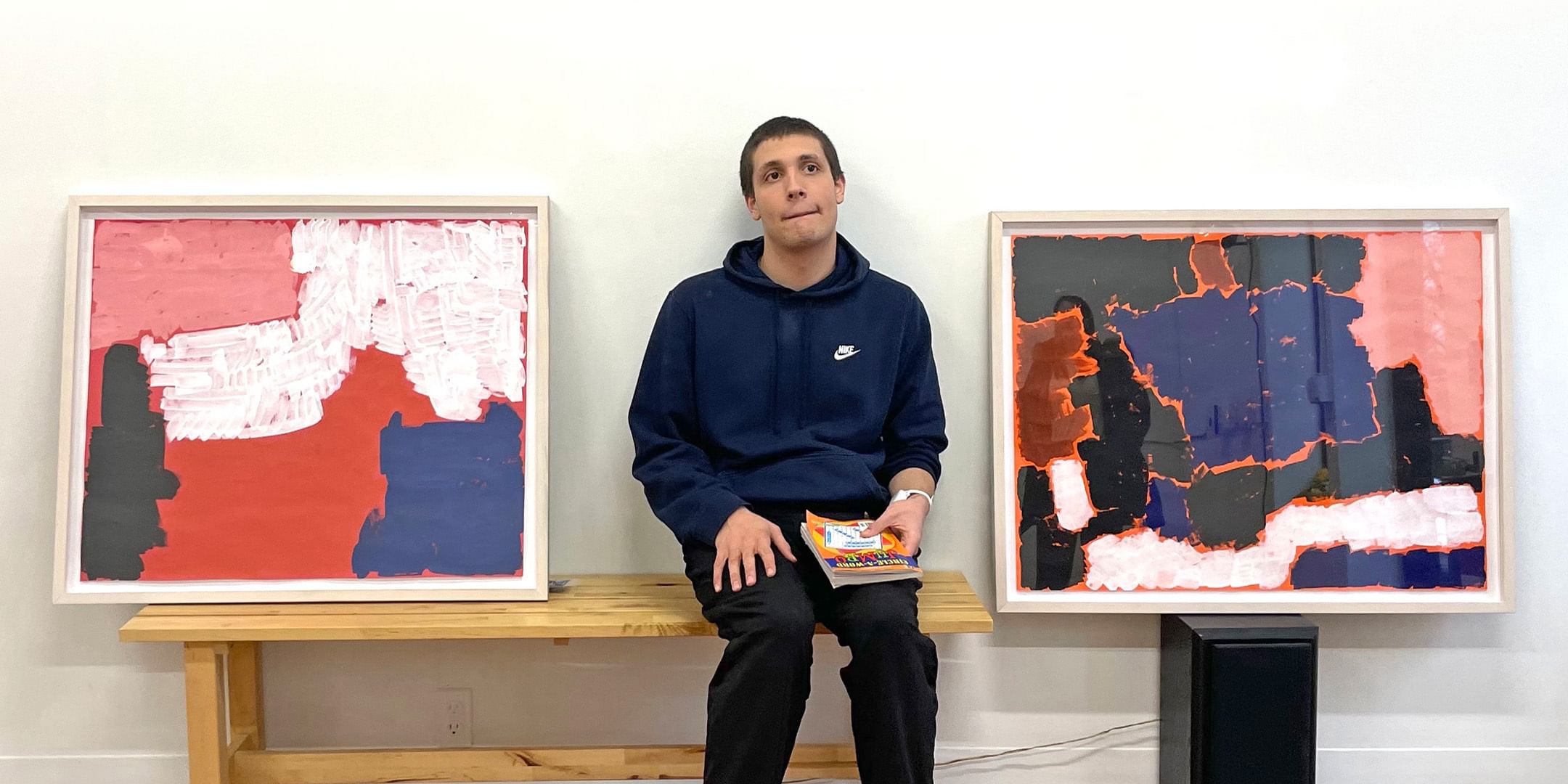 A young white man in a navy hoodie, holding a crossword puzzle book, sits between two abstract paintings.