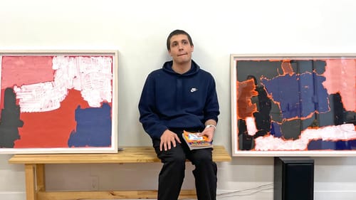 A young white man in a navy hoodie, holding a crossword puzzle book, sits between two abstract paintings.