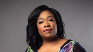 Cann, a Black woman, poses with one hand on her hip, wearing a flowing black and floral-printed wrap shirt.