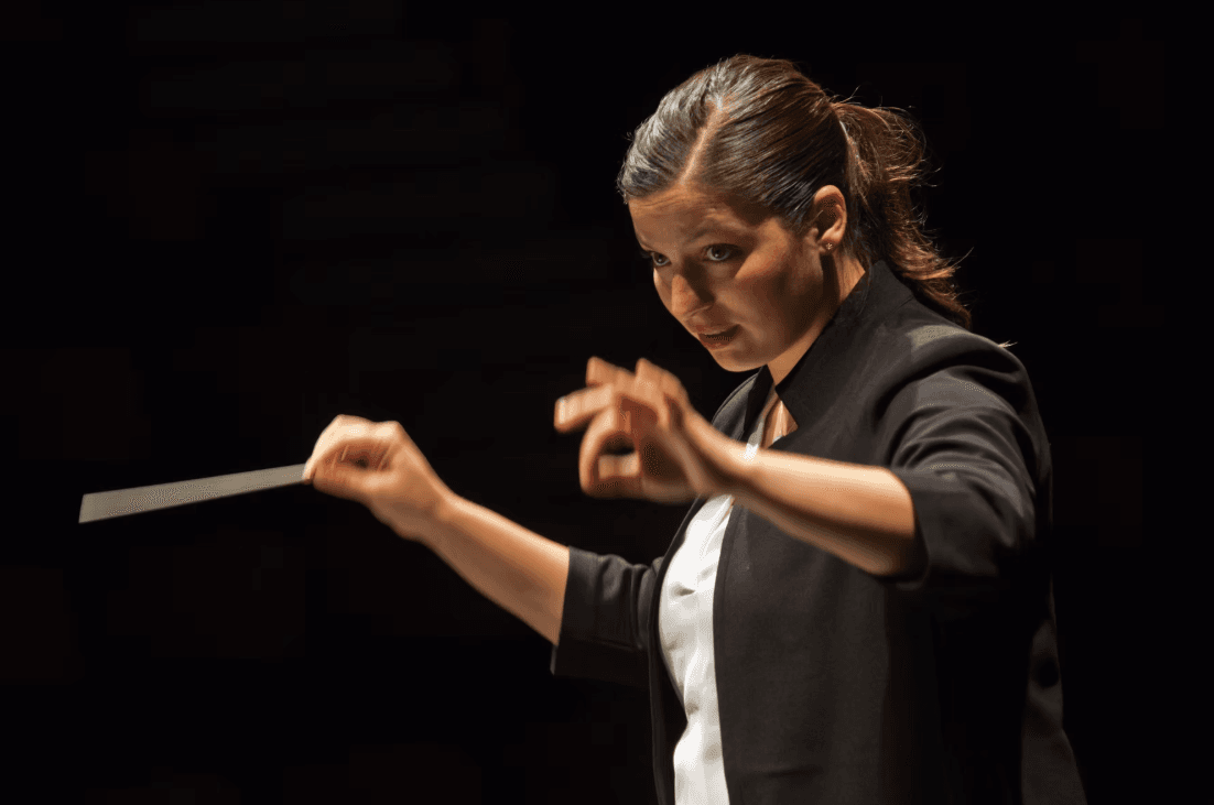 Closeup on Di Rossi, a white woman with brown hair in a ponytail and a black jacket, conducting with a baton.