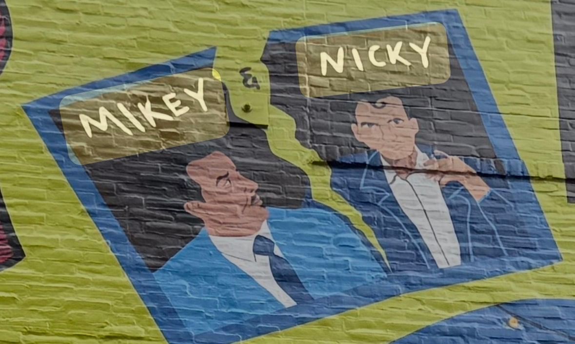 Closeup on a detail of a painted mural showing two men in blue suits appearing in a picture that’s ripping apart.