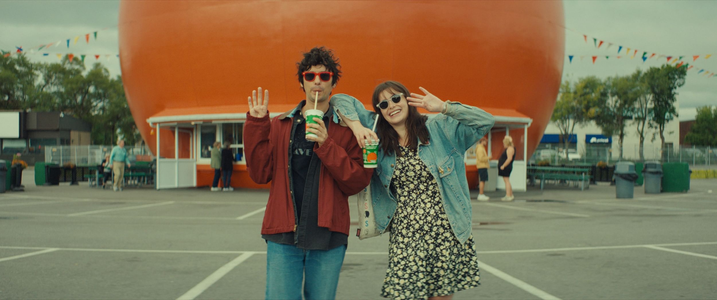 A young man & woman, in casual clothes & sunglasses, smile & wave in front of a giant round orange smoothie stand.