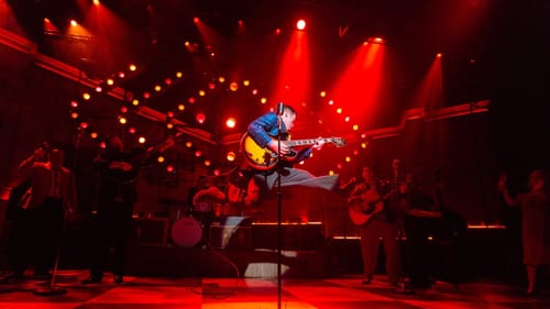 Actor Wren leaps into the air with a kick split while playing guitar in a red-tinted stage with other performers.