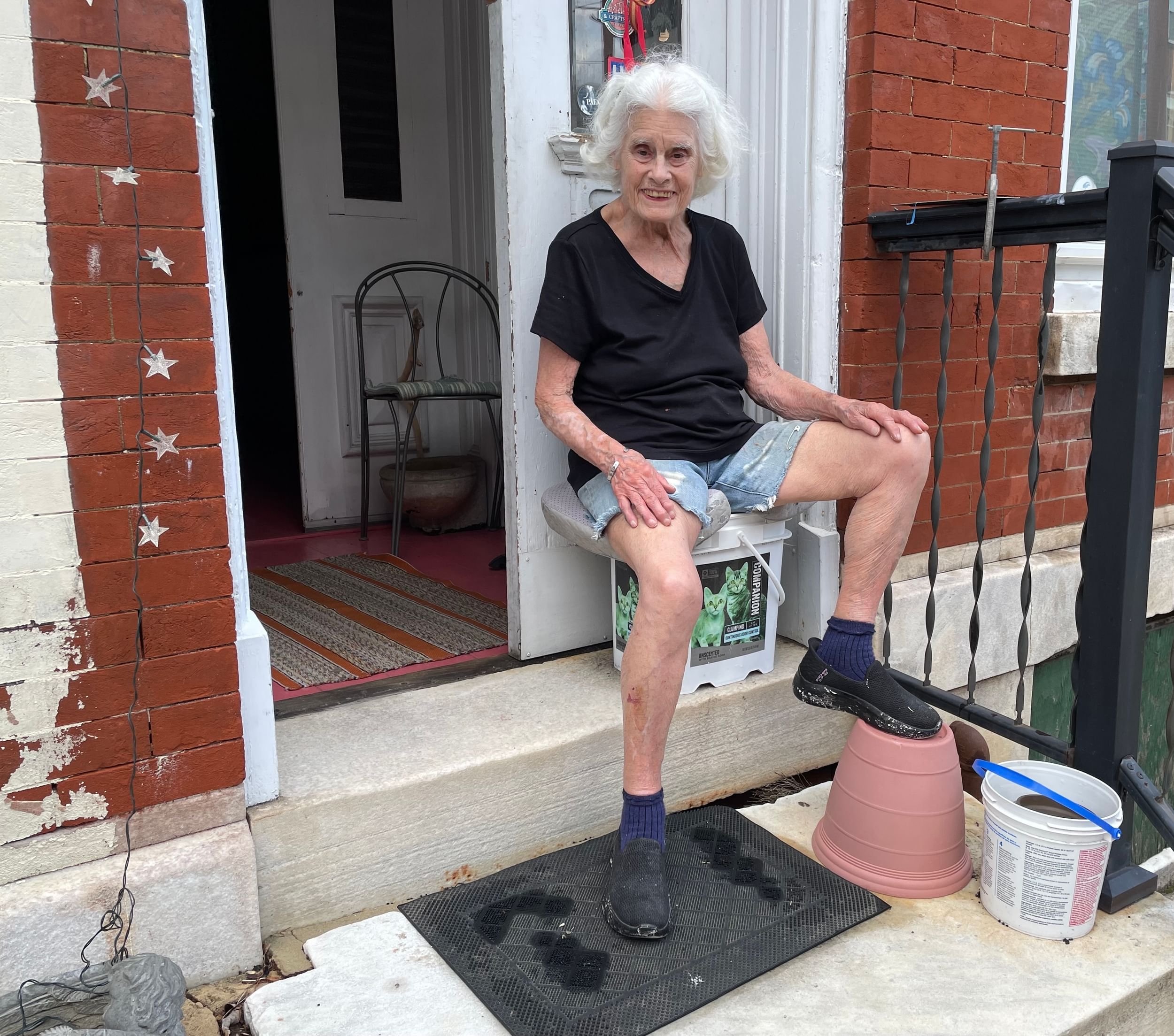 Minich in a plain black T and shorts sits on her stoop on a bucket, her foot on a planter. The front door open behind her