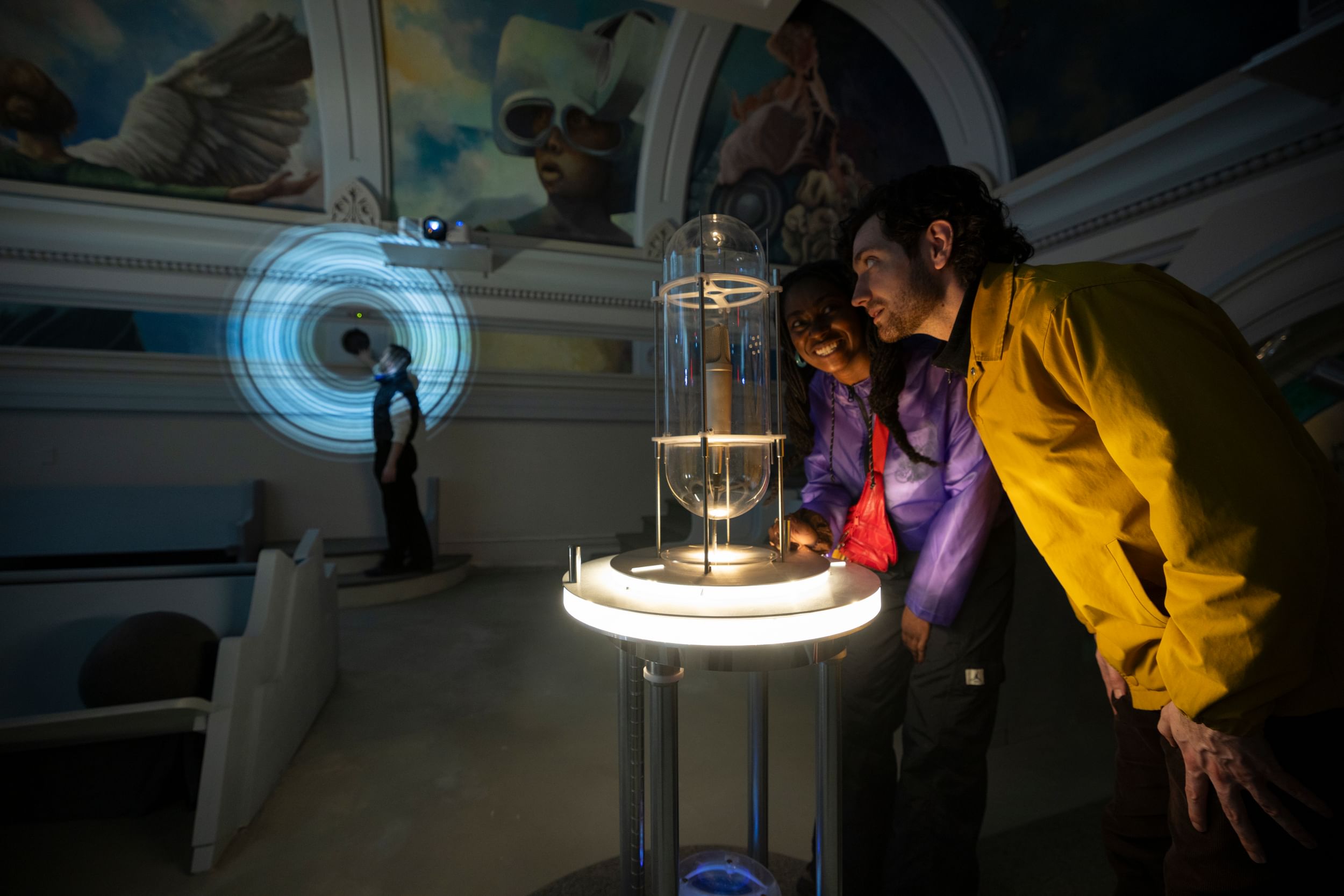 In a darkened room with mural-decorated vaulted ceilings, two people look animatedly into a glass contraption holding a mic.