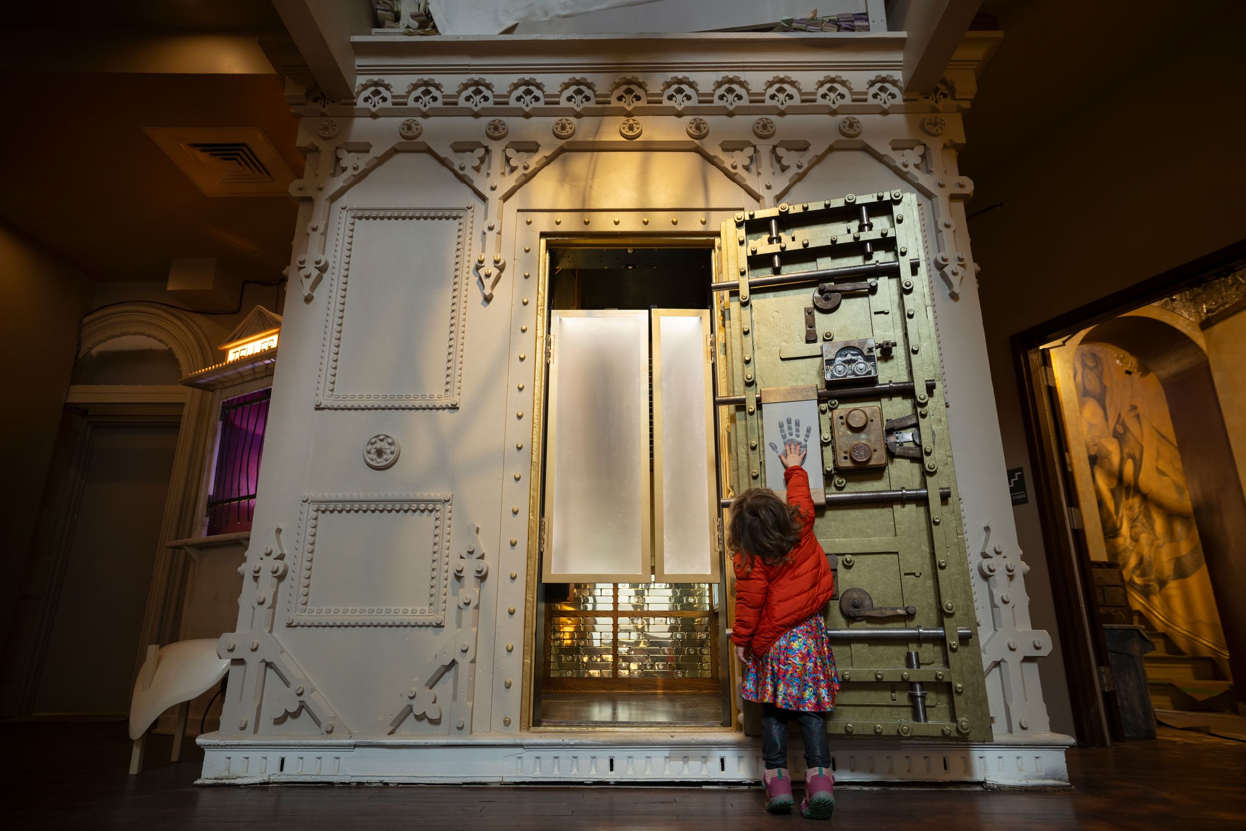 A little girl reaches up to a rectangle with a handprint sensor on it, outside a large 19th-century vault full of gold bars