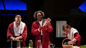 A scene from Minor Character: three actors, wearing large red cardigans, seem to be prepping dinner on a long wooden counter