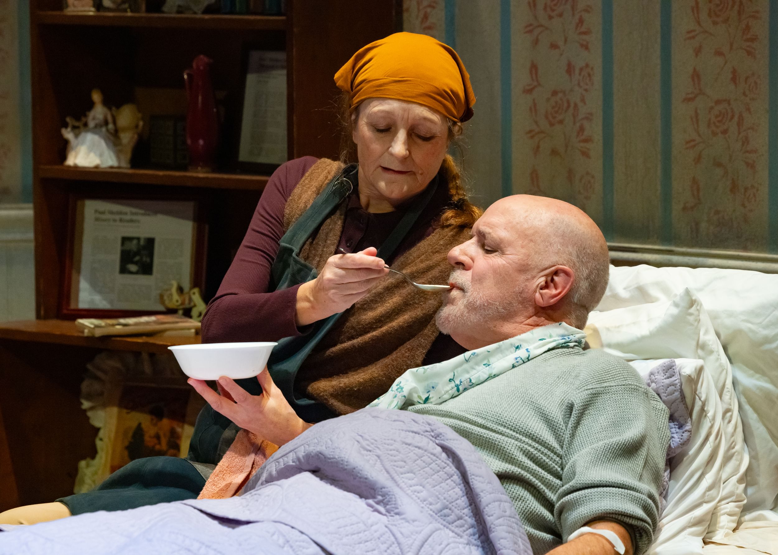 Gibson, in an orange bandana, tenderly spoons soup into Graham’s mouth as he lies flat in bed.