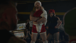 Two wrestlers, one in front with his arms crossed, and one behind him stands with her arms at her sides