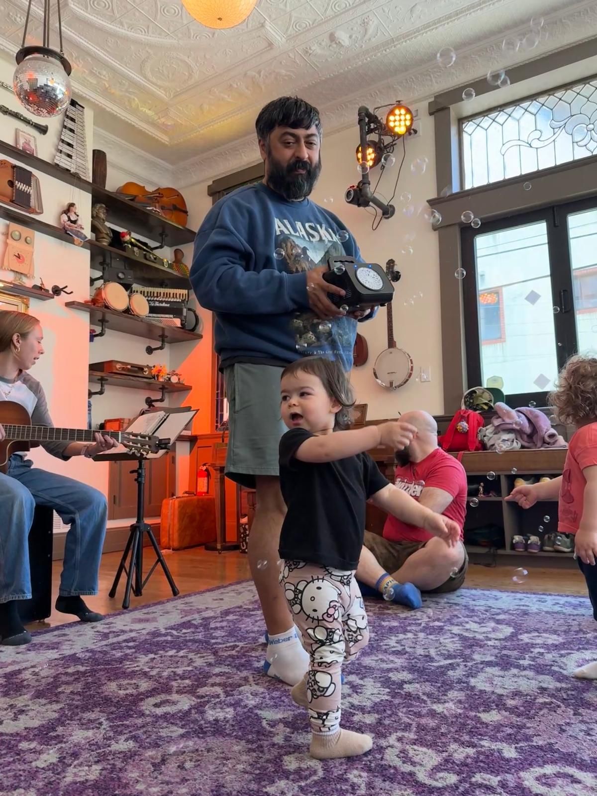 In a colorful room, Neil stands on a purple carpet holding a small bubble machine while his toddler dances in the bubbles.
