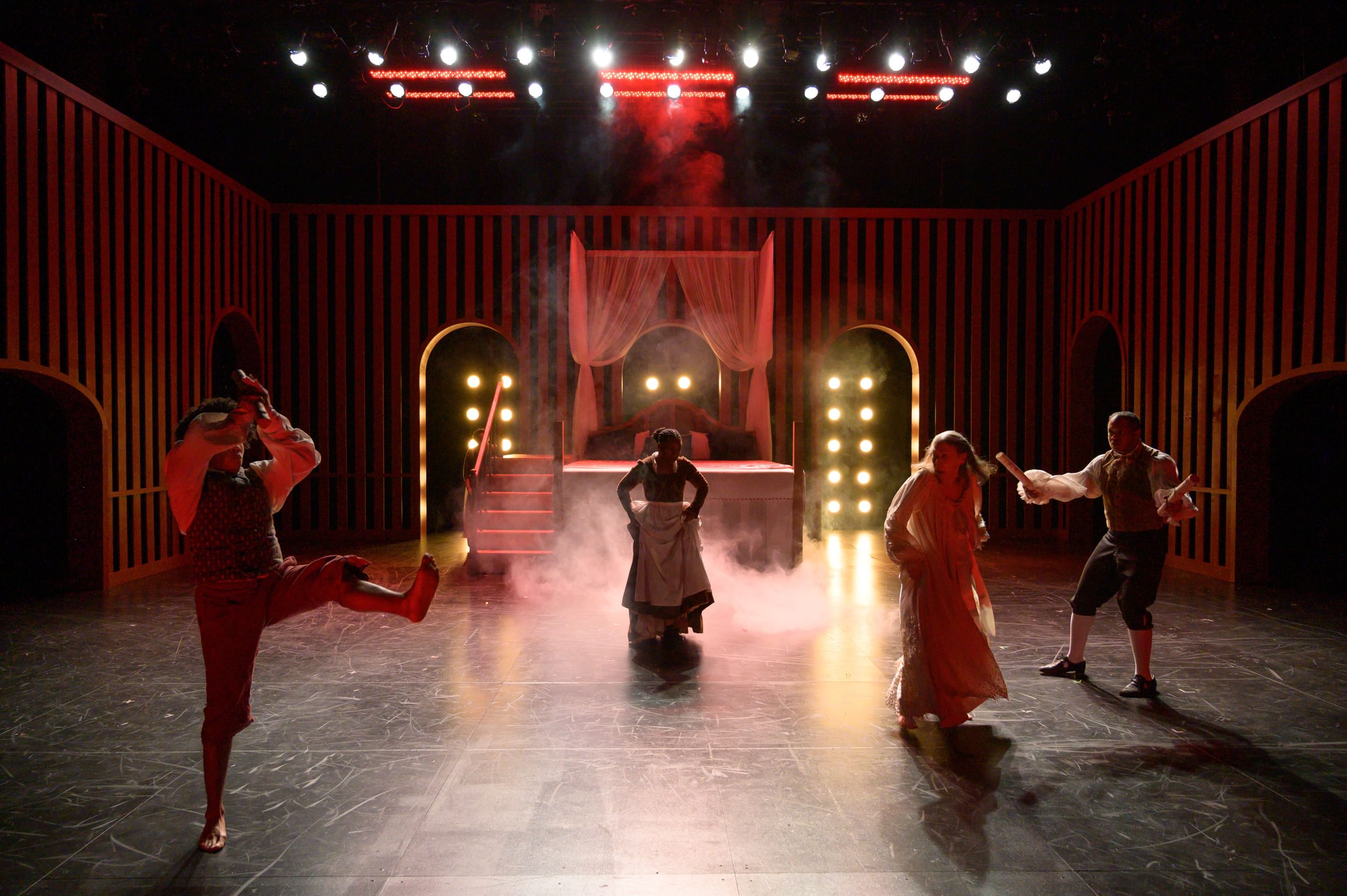 On a surreal stage with red striped walls, rows of yellow lights, and fog, actors cavort in front of a large curtained bed