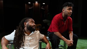 Grant, a Black man with long locs, lounges on a bench with Jimeniz, a worried-looking Latino man wearing a red shirt.