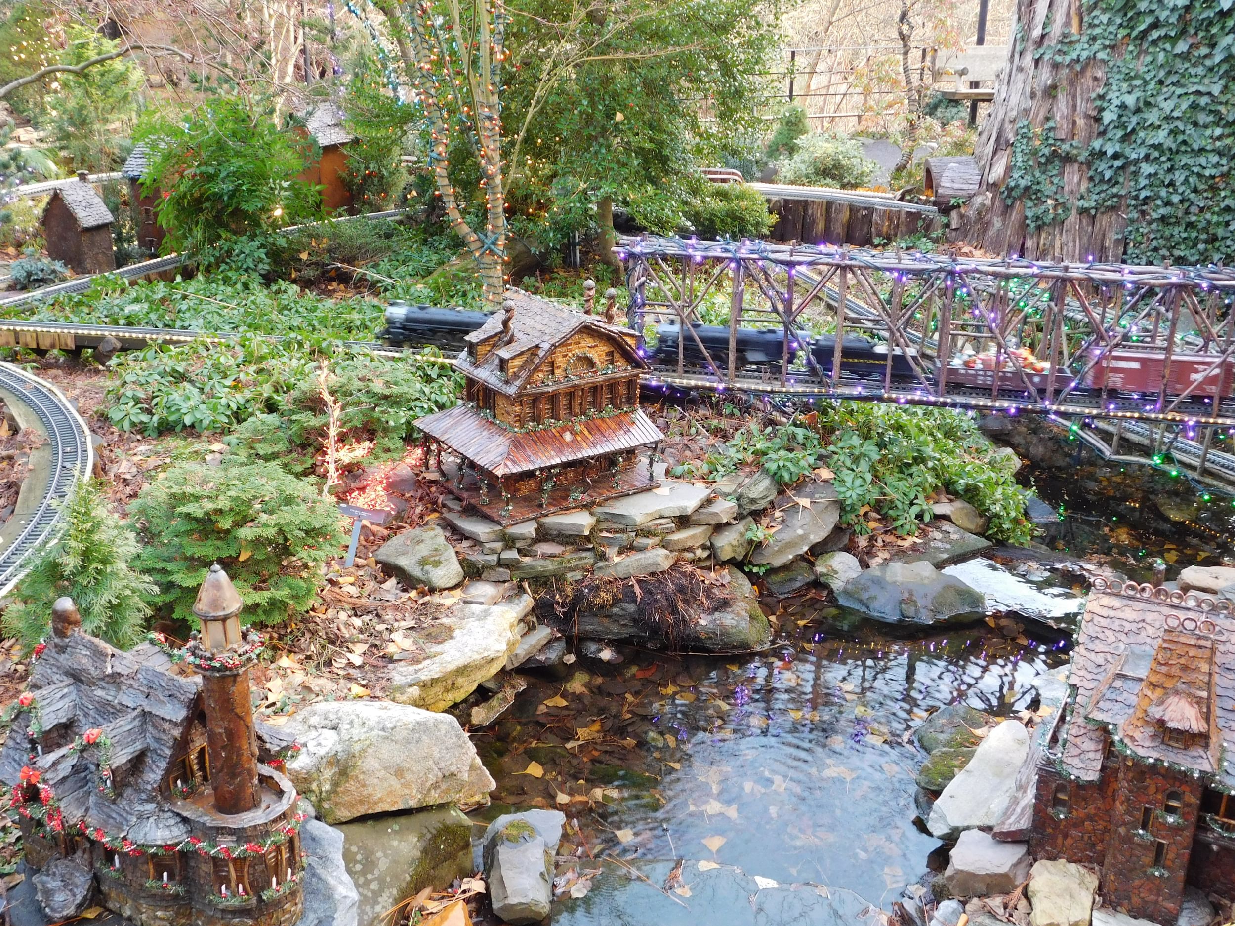 A model railway stretches over a small bed of water, a model train station structured at the opposite end in a garden area