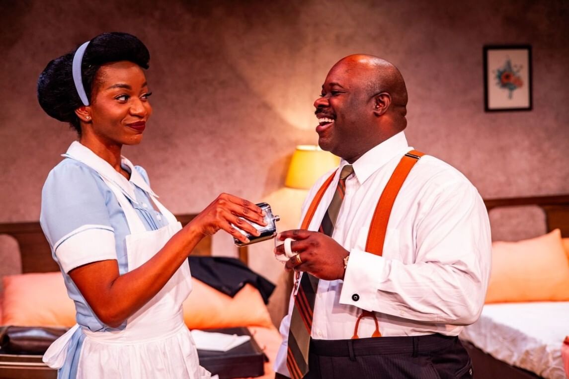 Nixon, in a maid’s uniform, flirtatiously pours a flask into Davis’s coffee cup while he laughs. They are in a hotel room.