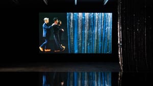 In a dark room, a large screen shows 3 people walking toward a glittering metallic curtain, all reflected on the shiny floor