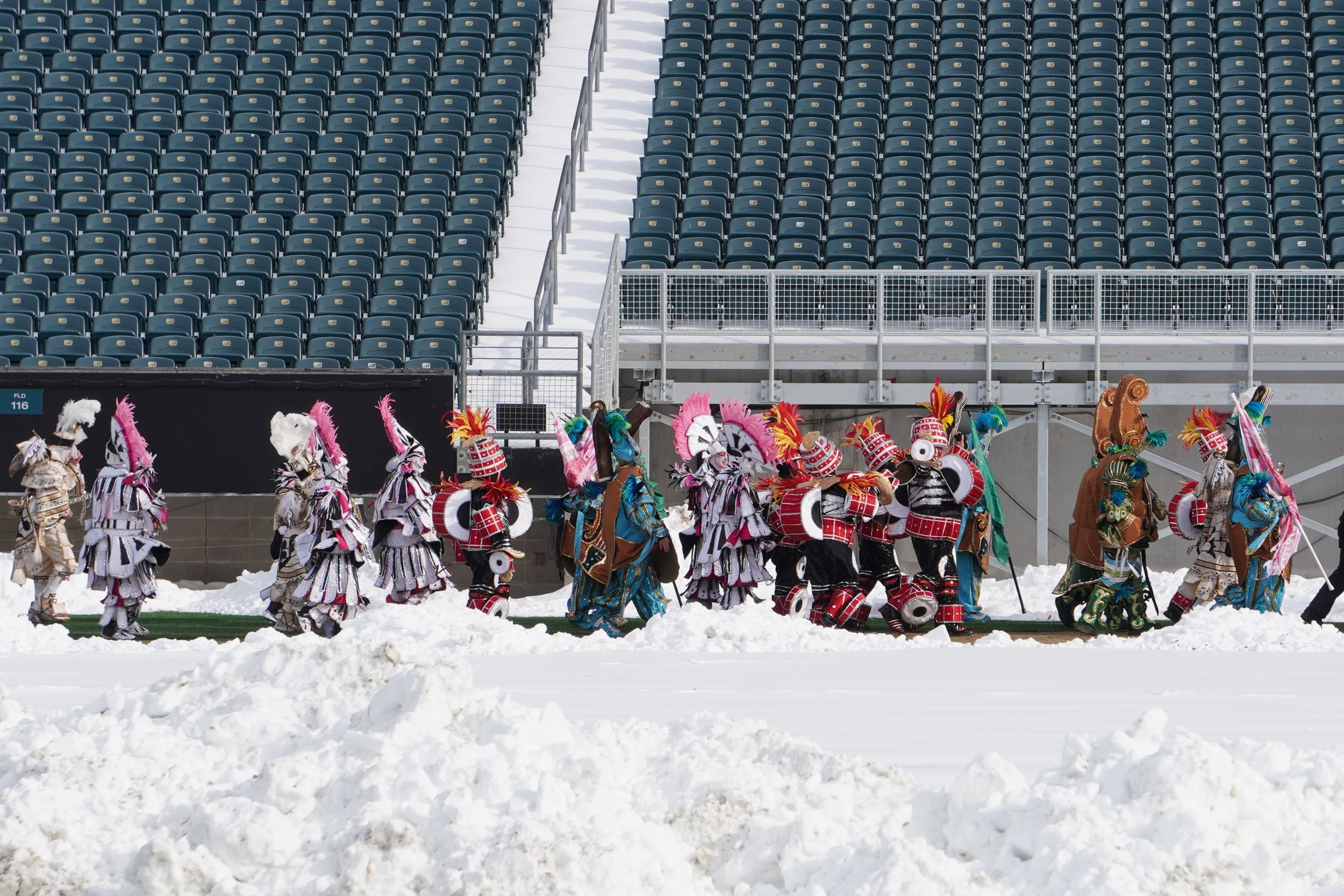 Mummers in opulent, colorful feathered costumes seen over piles of snow in the foreground, with empty stadium seats behind