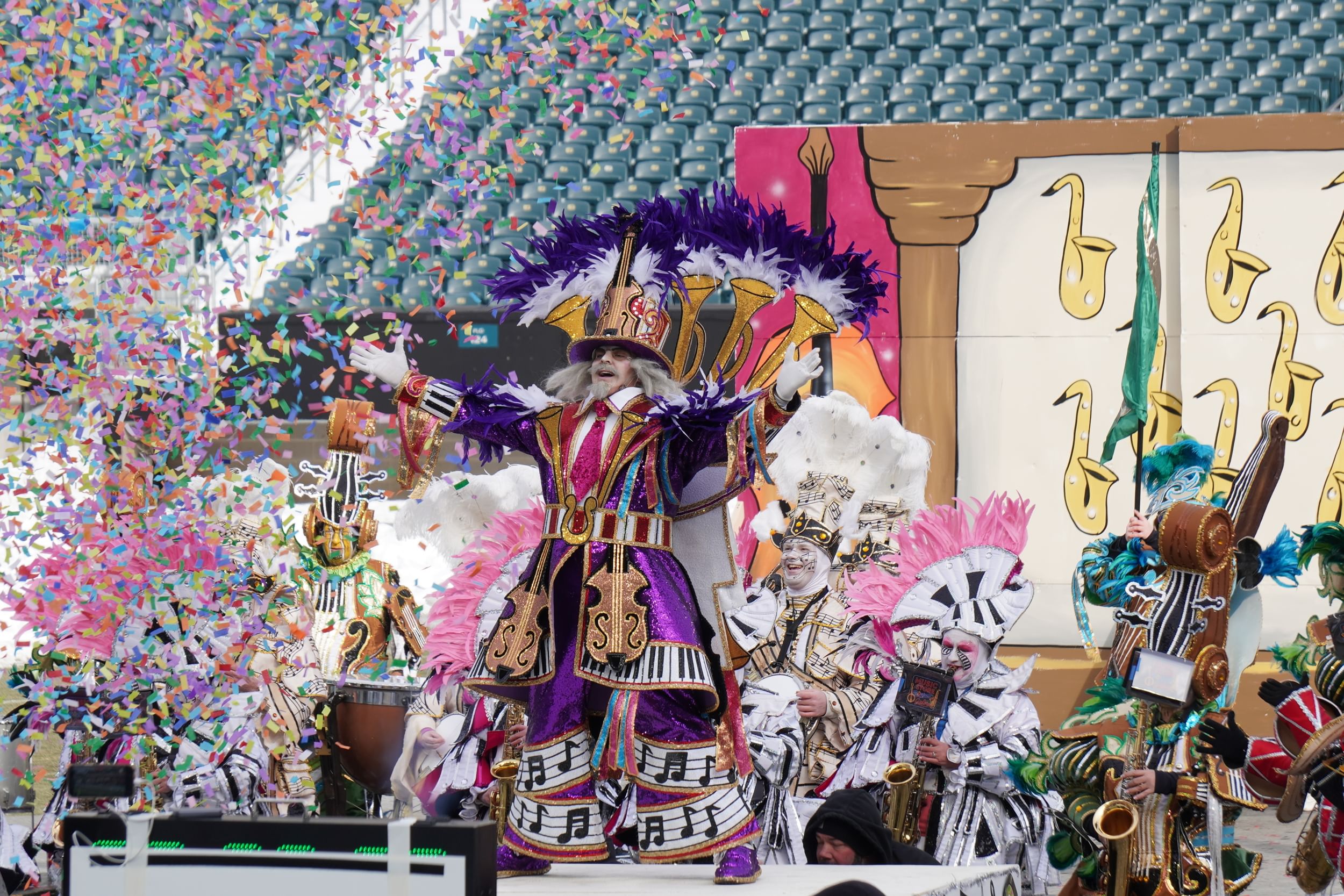 A man in an outlandish purple & gold sequined feathered costume spreads his arms as costumed band plays behind under confetti