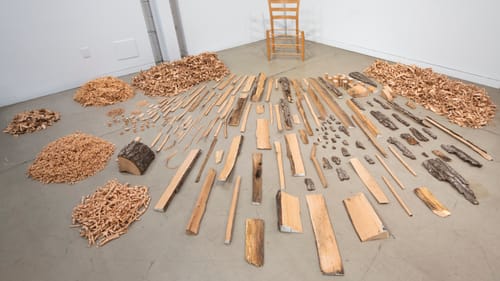 Art piece described above, set in a white-walled gallery corner on a well-worn gray floor. The wooden chair slender and pale.