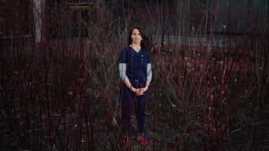 A woman nurse with pale skin and brown hair, wearing blue scrubs, looks calmly outward from a thicket of red spring branches