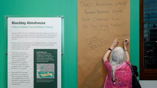 Seen from behind, a woman with a long gray braid writes on a roll of brown butcher paper next to museum signage.