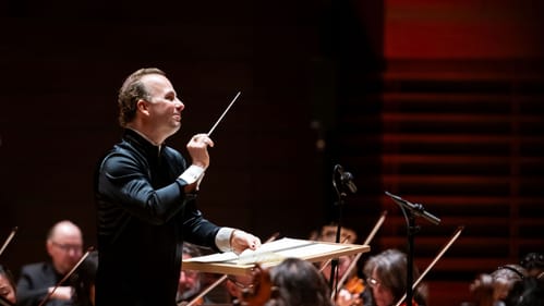 Closeup on Nézet-Séguin while he conducts, smiling passionately in a silky black shirt with large white cuffs.