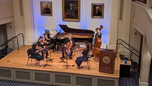 View from mezzanine above of six diverse musicians playing on a small stage under founding father portraits.