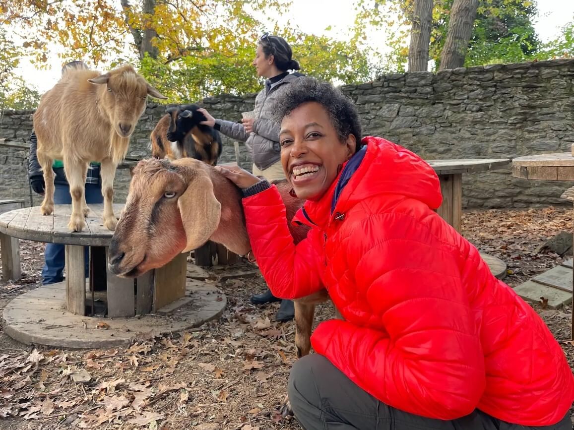 Francis, a Black woman wearing a red coat, smiles widely while petting a friendly brown goat in an outdoor enclosure.